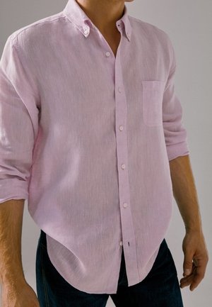 Uomo che indossa una camicia rosa chiaro a manica lunga con polsini arrotolati e pantaloni scuri su uno sfondo semplice.