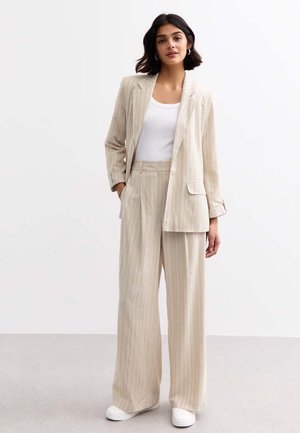 Costume rayé beige avec un blazer ajusté et un pantalon à jambes larges, en tissu léger. Associé à un haut blanc et des baskets blanches.
