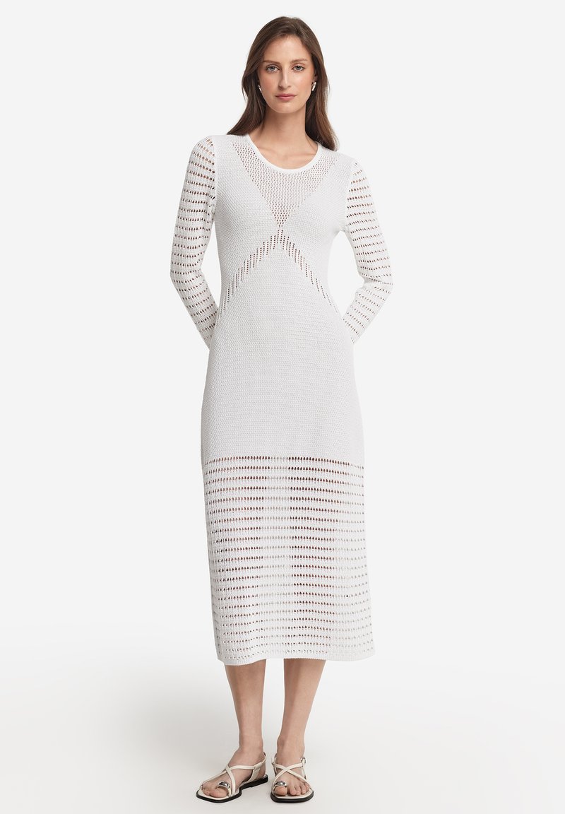 Weißes Strickkleid mit langen Ärmeln, das geometrische Ausschnitte und eine gerippte Textur aufweist, bis zur Mitte der Waden reicht und mit flachen Sandalen kombiniert ist.
