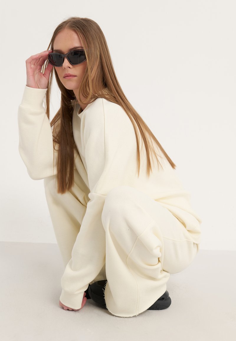 Felpa crema oversize e pantaloni larghi, tessuto testurizzato; occhiali da sole neri; modella accovacciata su uno sfondo neutro.