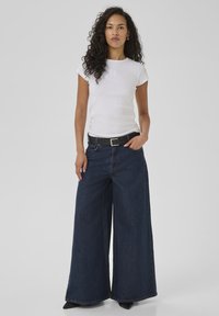 Woman with curly hair wearing a fitted white t-shirt, wide-leg dark blue jeans, black belt, and black pointed shoes, standing against a gray background.