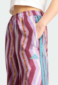 Calças desportivas multicoloridas com padrões em zigzag em roxo, castanho-escuro e azul-turquesa, com texto amarelo, uma cintura elástica e bolsos laterais.