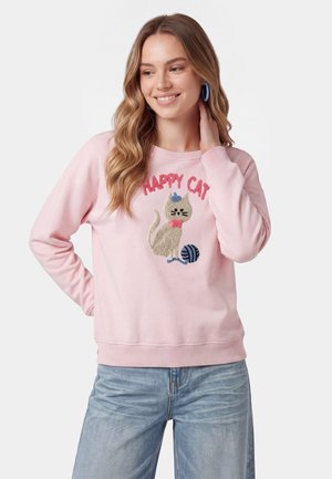 Kvinde med langt, bølget hår iført en pink sweatshirt med et motiv af en kat og garn samt teksten "HAPPY CAT," sammen med blå jeans.