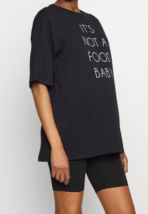 Osoba nosząca czarny, oversize'owy T-shirt z napisem „TO NIE BRZUSZEK PO JEDZENIU” oraz czarne dopasowane szorty na jednolitym białym tle.