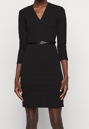 Femme portant une robe noire ajustée avec des manches trois-quarts et une fine ceinture noire à la taille.