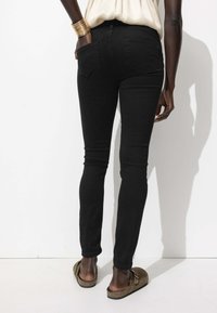 Jean skinny noir avec une texture lisse, taille mi-haute et deux poches arrière. Porté avec des sandales à enfiler de couleur terre.