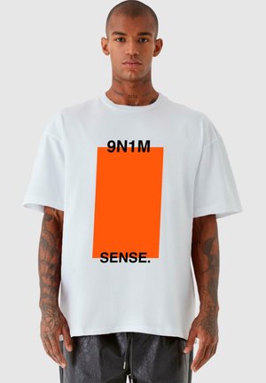 Hvit oversized t-skjorte laget av bomull, med et oransje rektangulært grafikk og svart tekst som leser "9N1M SENSE." på forsiden.