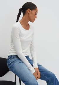 Vrouw met gevlochten haar, gekleed in een wit shirt met lange mouwen en een blauwe spijkerbroek, die zijwaarts op een zwarte stoel zit tegen een effen achtergrond.