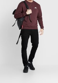 Sudadera burdeos con logo, combinada con pantalones vaqueros negros y zapatillas negras. Los accesorios incluyen una mochila negra con correas ajustables.