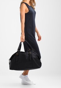 Borsa duffel nera con manici doppi, tasche laterali e accessori dorati, realizzata in un materiale testurizzato. Indossata da una persona in un vestito blu navy.