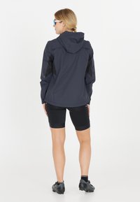 Lichte, waterdichte donkergrijze jas met een capuchon, zwarte accenten en ademende stof, gecombineerd met zwarte fietsshorts en schoenen.