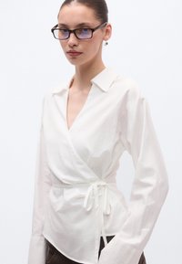 Άσπρο μπλουζάκι τύπου wrap από απαλό βαμβάκι, με βαθύ V-λαιμό, μακριά μανίκια και ζώνη στη μέση. Διαθέτει δομημένο γιακά και ροή στο σχήμα.