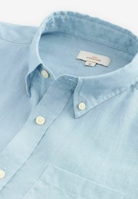 Camisa de lino azul claro con cuello abotonado, botones blancos, un bolsillo en el pecho y una etiqueta que dice "NEXT SIGNATURE M".