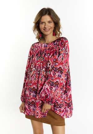 Løsbluse i levende pink, rød og sort abstrakt mønster med ballonsærmer og en gathered halsudskæring, parret med en brun nederdel.