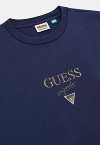 Navyblauwe Guess Originals t-shirt met gouden logo en een driehoekig panterprint-embleem onder de tekst nabij de halslijn.
