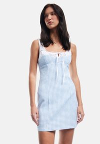 Lyseblå gingham kjole med tætsiddende bodice, hvid flæset halsudskæring og bundet frontdetalje. Lavet af blødt, struktureret stof.