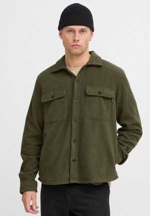 Olijfgroene fleece shirtjack met twee borstzakken, zwarte knopen en een kraag. Model draagt een zwarte beanie en een lichtgekleurde shirt eronder.
