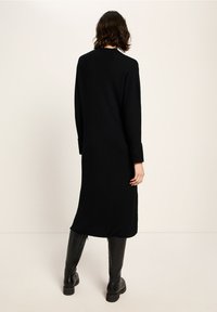 Person mit schulterlangem braunem Haar, die ein langes schwarzes Rippstrickkleid und kniehohe schwarze Stiefel trägt, mit dem Rücken zu einer einfarbigen Hintergrund.