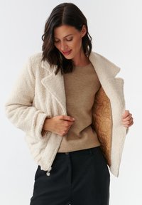 Crème, fluffy fleece jacket met een uitgesneden kraag, ritssluiting aan de voorkant en een lichtbruine gevoerde binnenkant. Model draagt een beige trui en zwarte broek.