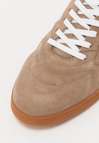 Braune Wildledersneaker mit weißen Schnürsenkeln. Mit gesticktem Wellenmuster, runder Zehenform und strukturiertem Gummisohle in Beige. Glatte Oberflächenstruktur.