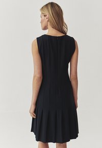 Ärmelloses schwarzes Kleid mit einem gefalteten Rock, ausgestattet mit einem subtilen Reißverschluss auf der Rückseite und glattem Stoff, der für eine taillierte Silhouette sorgt.