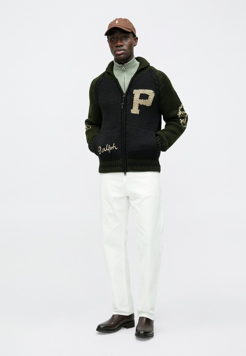 Cardigan tricoté en vert foncé et noir, avec un grand "P" beige et des détails brodés "Ralph", associé à un pantalon blanc et des bottes marron.