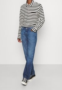 Camisa polo de manga larga a rayas en blanco y negro, combinada con jeans de mezclilla azules y zapatillas negras. Presenta un bolsillo en el pecho y un ajuste relajado.