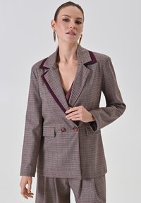 Bruine geruite blazer met contrasterende bordeauxrode revers en knopen. Tailored fit, met een diepe V-hals en twee voorzakken.