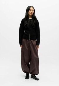 Pull à fermeture éclair noir avec manches longues, associé à un pantalon marron brillant surdimensionné avec des poignets élastiques. Des chaussures noires sans lacets complètent la tenue.