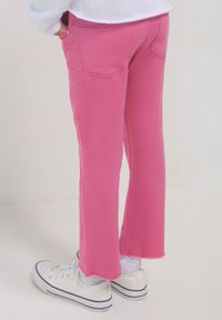 Person, die leuchtend pinkfarbene, verkürzte Jeans mit rohem Saum, weiße Socken, weiße Low-Top-Sneaker und ein weißes Oberteil trägt, mit Händen in den Taschen.