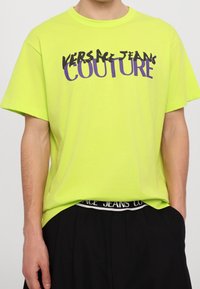 Neon zelené bavlněné tričko s krátkým rukávem, které má černý a fialový grafický text s nápisem "VERSACE JEANS COUTURE."