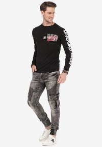 Chemise noire à manches longues avec imprimé graphique, associée à un pantalon cargo gris usé avec des accents texturés et des poignets élastiques. Baskets blanches.