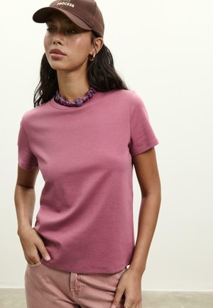 Femme portant un t-shirt mauve, un pantalon rose clair, un collier en perles violettes et une casquette marron avec le texte "THE PROCESS", se tenant contre un fond uni.