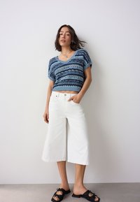 Blauer, kurz geschnittener Strickpullover mit strukturierten Streifen, kombiniert mit weißen, weit geschnittenen Hosen und schwarzen Pantolette-Sandalen. Natürlich wirkende Umgebung.