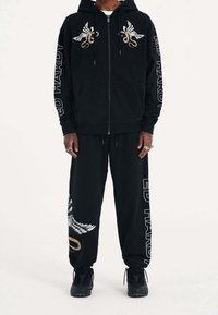 Svart zip-up hoodie och sweatpants med grafik av vingar och orm, med "ED HARDY" text längs ärmarna och byxorna, tillverkad av mjukt tyg.