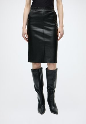 Femme portant une jupe crayon en cuir noire arrivant aux genoux et des bottes en cuir noires montant jusqu'aux genoux, debout devant un fond uni.