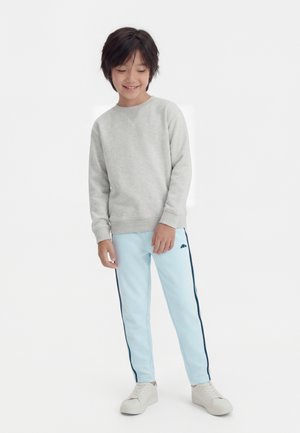 Niño sonriente con sudadera gris clara, pantalones deportivos azul claro con franjas oscuras en los lados y zapatillas blancas, posando frente a un fondo liso.
