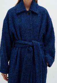 Kvinna iklädd en strukturerad, mörkblå och svart, bältesjacka med stor krage och en svart knapp, med händerna delvis i fickorna.