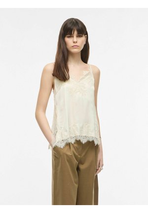 Femme aux cheveux bruns et raides portant un haut sans manches crème orné de dentelle et un pantalon marron ample, debout avec une main dans la poche.