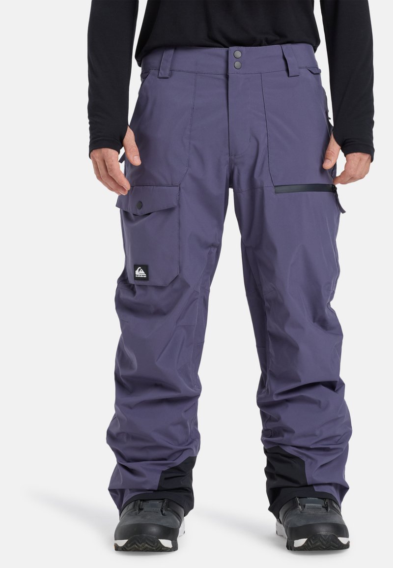 Pantalones de nieve morados, resistentes al agua, con múltiples bolsillos cargo, cintura ajustable y puños reforzados. Presentan un parche con el logo y detalles con cremalleras.