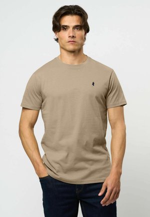 Άνδρας με σκούρα μαλλιά φοράει απλό μπεζ T-shirt με στρογγυλή λαιμόκοψη και μικρό σκούρο λογότυπο στο στήθος, σε συνδυασμό με σκούρα μπλε τζιν, στέκεται μπροστά σε απλό φόντο.