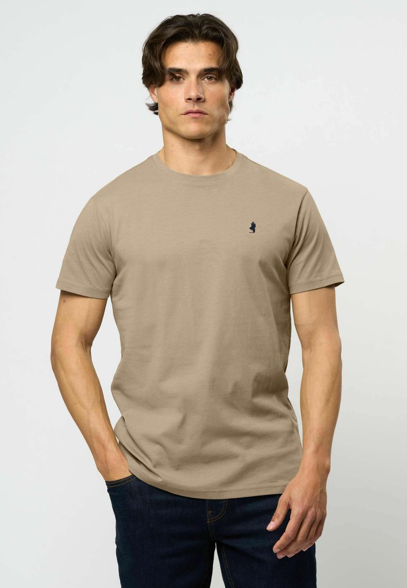 Άνδρας με σκούρα μαλλιά φοράει απλό μπεζ T-shirt με στρογγυλή λαιμόκοψη και μικρό σκούρο λογότυπο στο στήθος, σε συνδυασμό με σκούρα μπλε τζιν, στέκεται μπροστά σε απλό φόντο.