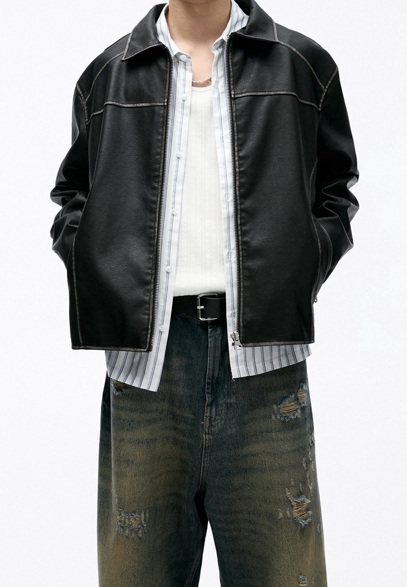 Black leather jacket with a collar, contrasting stitching, and zip closure, layered over a white striped shirt and distressed denim pants.