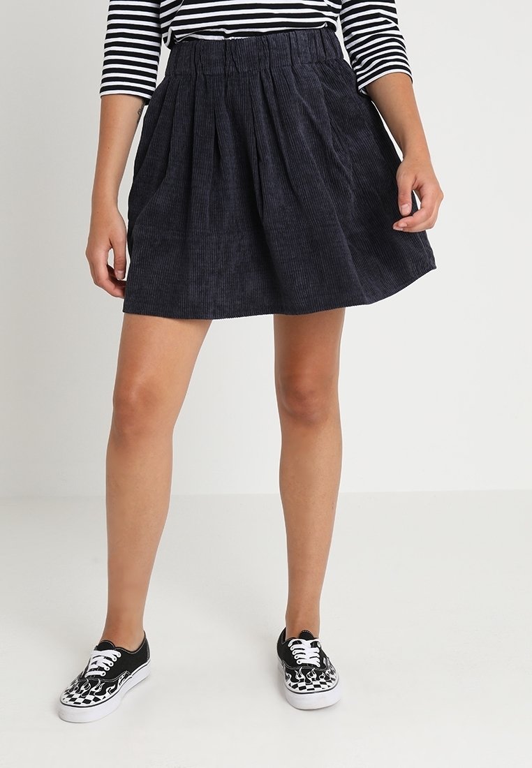 Femme portant une jupe plissée en velours côtelé noire, un haut rayé noir et blanc, et des baskets noires avec un motif de flammes blanches, debout sur un sol blanc.