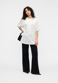 Λευκό υπερμεγέθες t-shirt με λαμπερό σχέδιο "G", σε συνδυασμό με μαύρο παντελόνι φαρδιάς γραμμής και μικρή μαύρη τσάντα. Λείο ύφασμα.