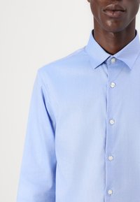 Vaaleansininen napillinen paita, jossa on klassinen kaulus, pitkät hihat ja sileä, tekstuuriinen kangas. Viisi nappia näkyvissä edessä.