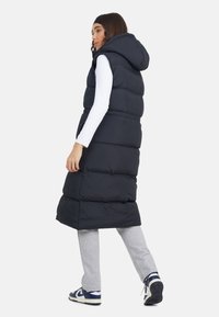 Długa, granatowa kamizelka puchowa z kapturem, posiadająca pikowany wzór i boczne kieszenie. Noszona na białej bluzce i szarych dresach.