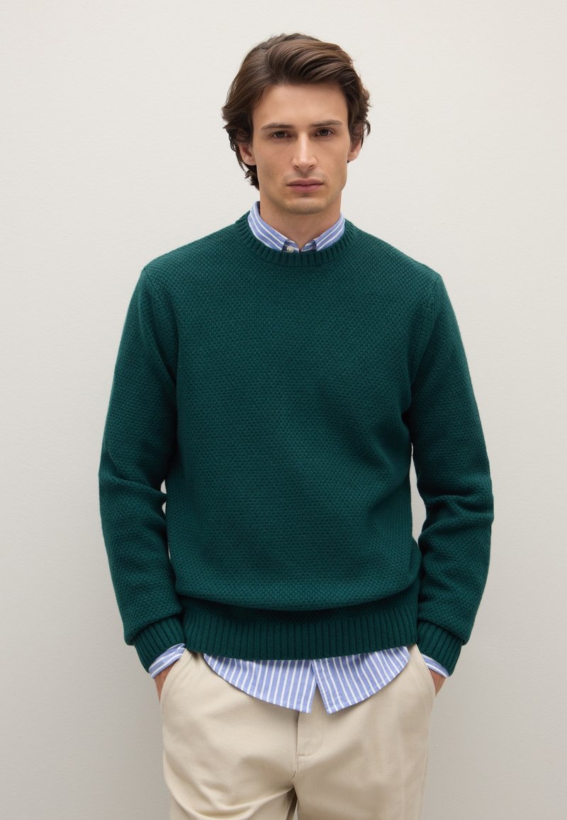 Dunkelgrüner, strukturierter Strickpullover mit Rundhalsausschnitt, darüber getragen ein hellblau-weiß gestreiftes Hemd mit Knöpfen und beigefarbene Hose.