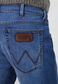 Vaqueros de denim azul con un parche de cuero marrón "Wrangler" en el bolsillo trasero, que presenta costuras naranjas y un diseño zigzag distintivo.