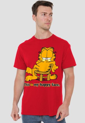 Red cotton t-shirt featuring a graphic of a sitting cartoon cat with a relaxed expression, and the text "This is my happy face." in black.